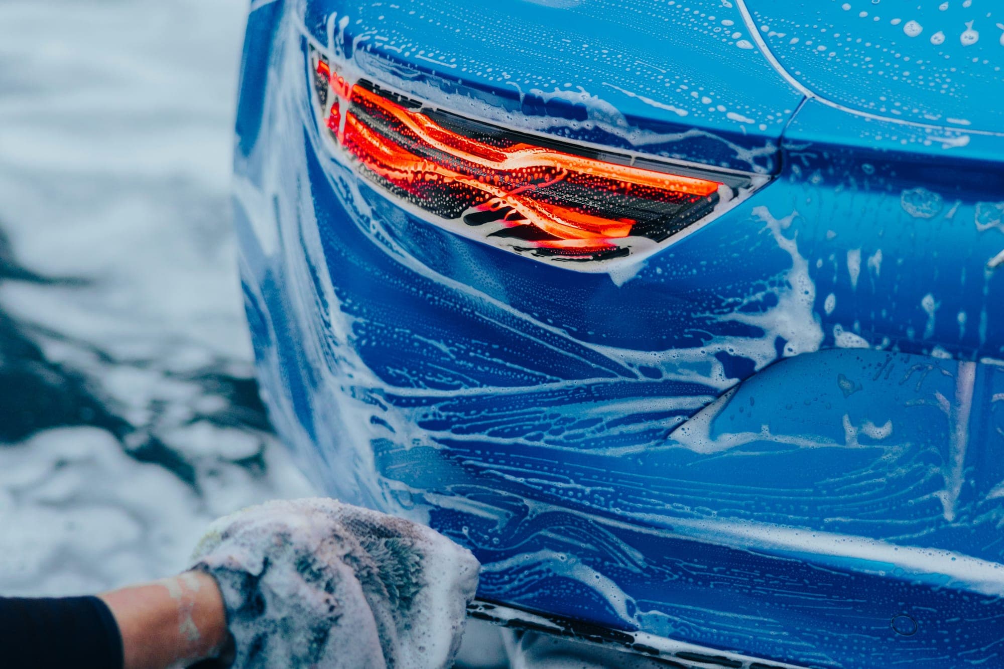 A gloved hand washing the soapy rear quarter of a blue car